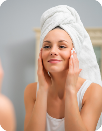 Woman with her hair in a towel touching her face in front of her bathroom mirror