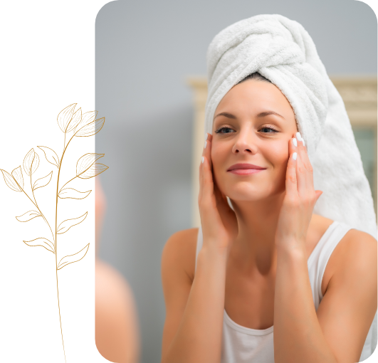 Woman with her hair in a towel touching her face in front of her bathroom mirror next to a decorative gold plant